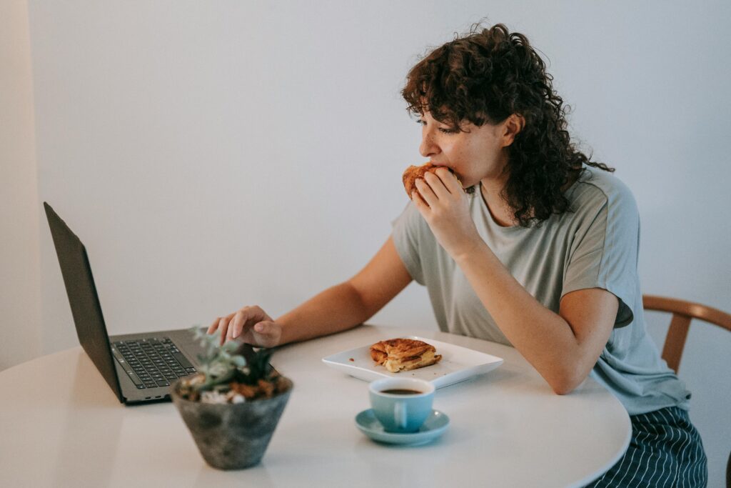 Young female with curly dark hair working with netbook while looking at screen and eating toasts with cup of coffee